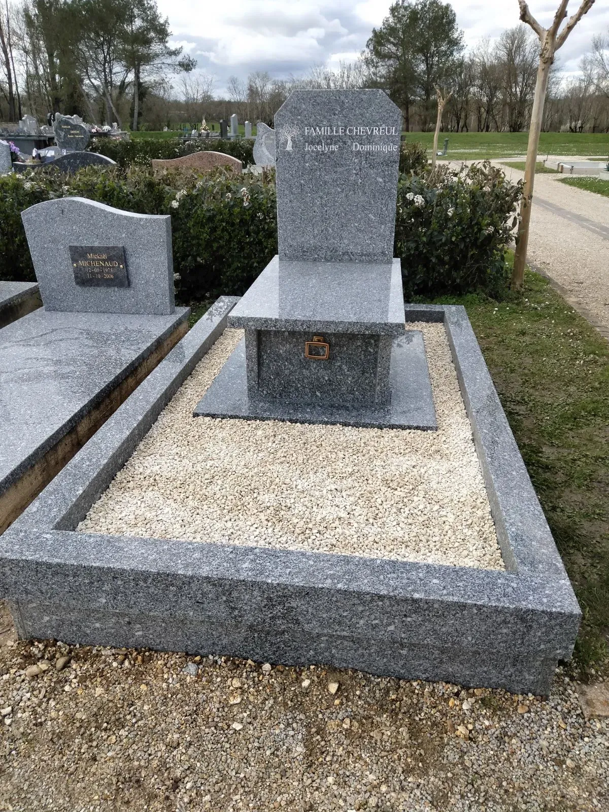 Sépulture en granit gris bordée d'un rectangle de gravier clair.